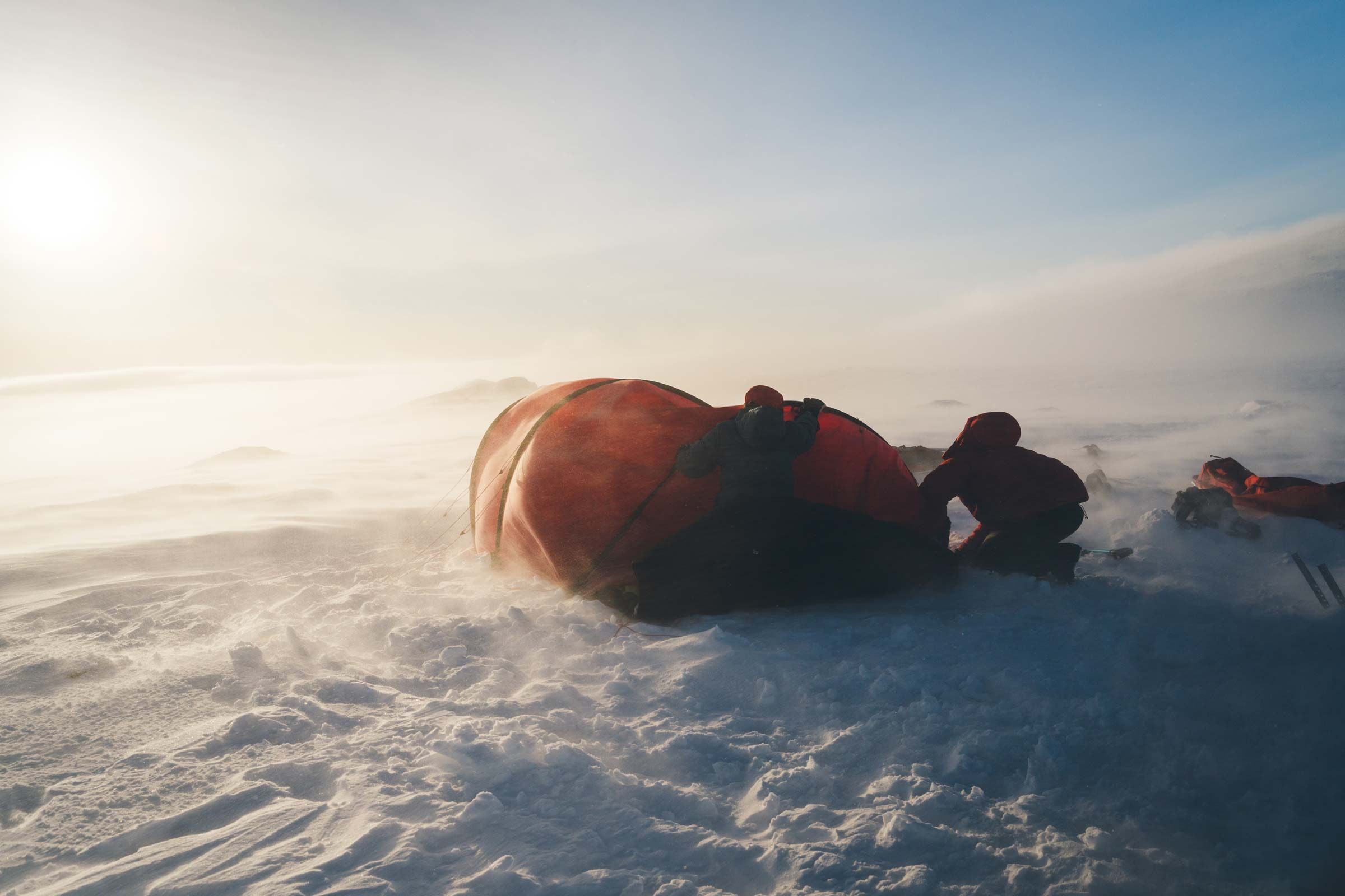 Retkeilijät ahkion vieressä tuiskussa matalan auringon valossa