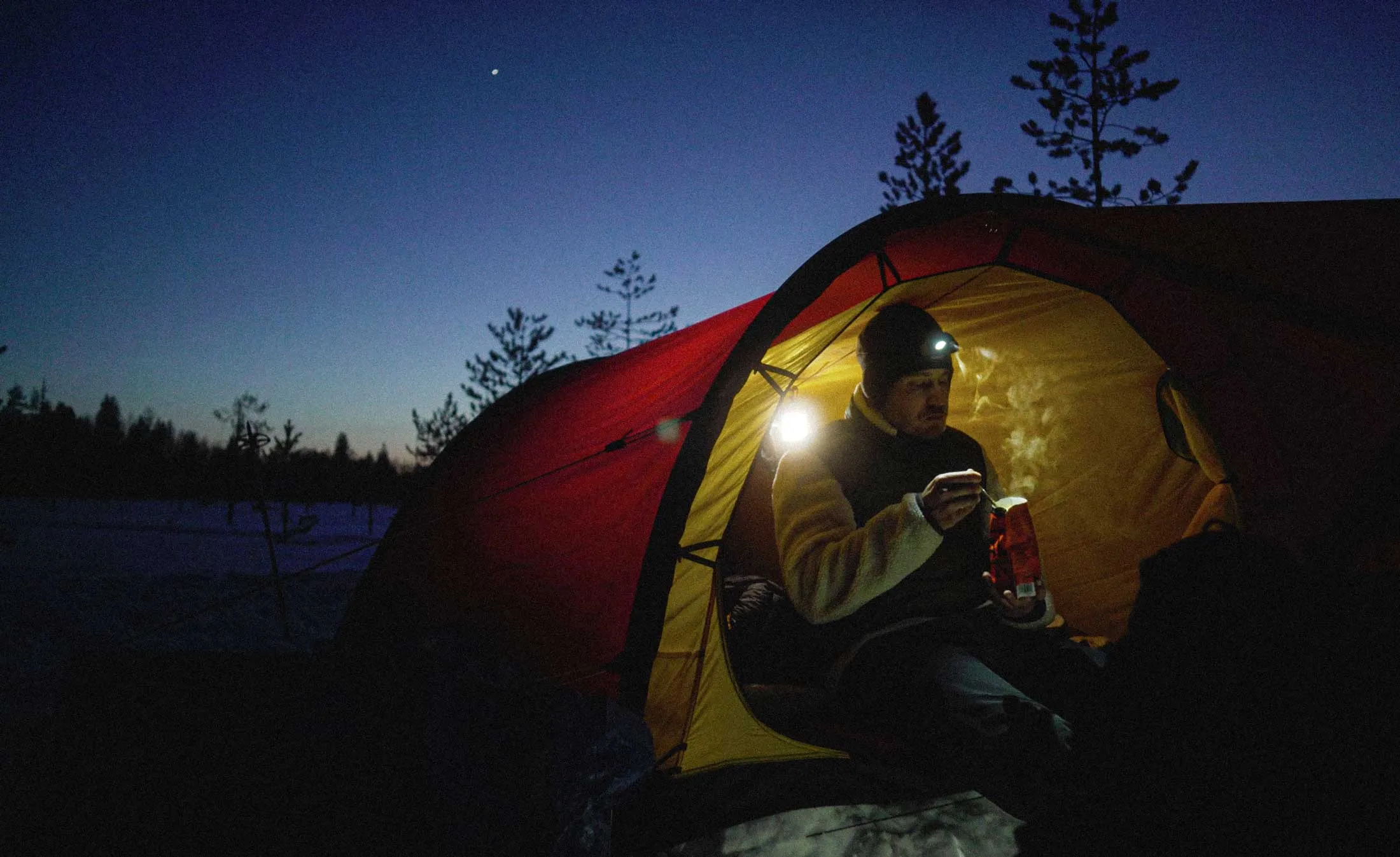 Retkeilijä istuu otsalamppu päässä valaistun teltan suulla iltahämärässä